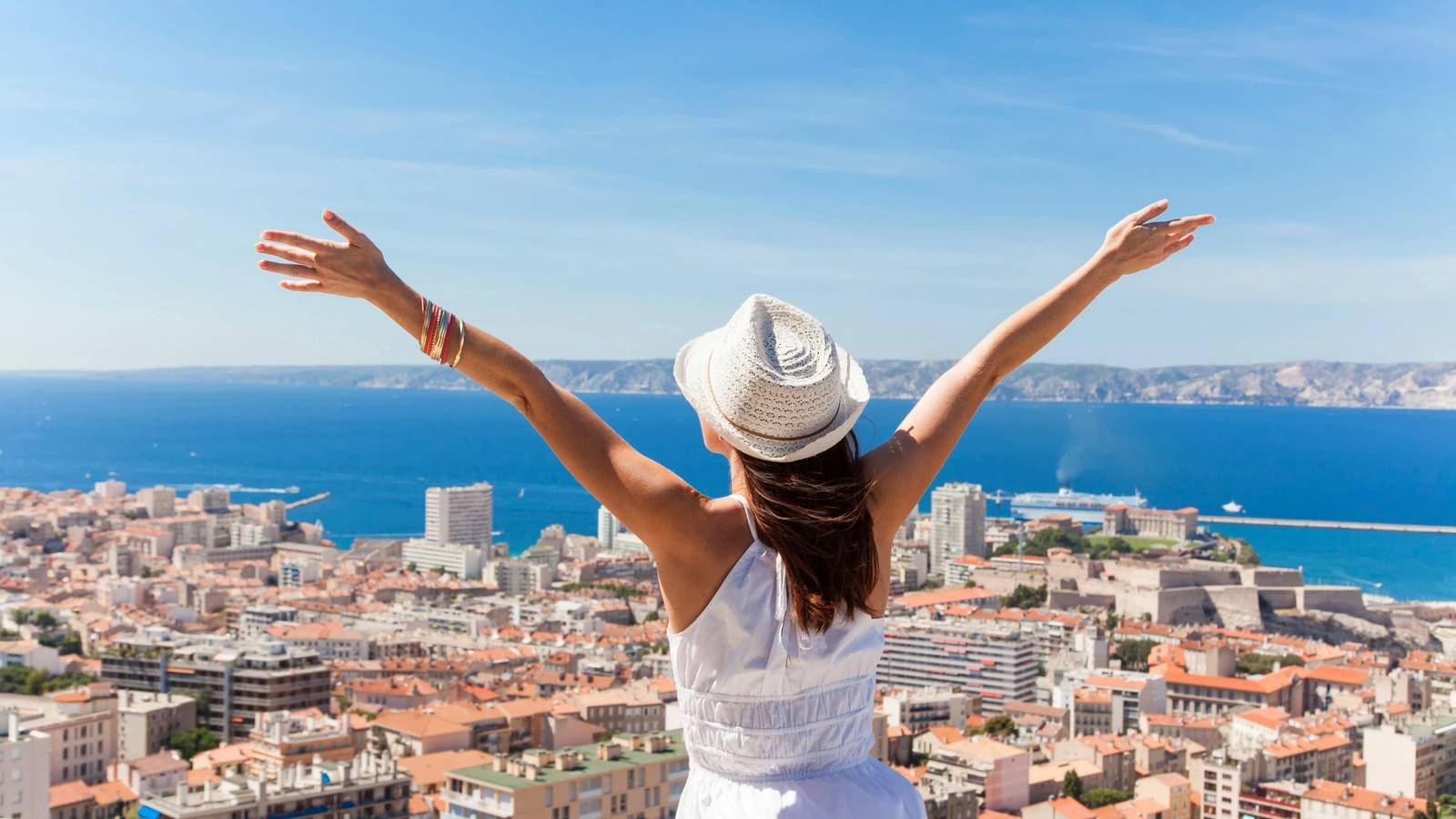 A woman in a white dress and hat stands with arms outstretched, overlooking a coastal city and blue sea.