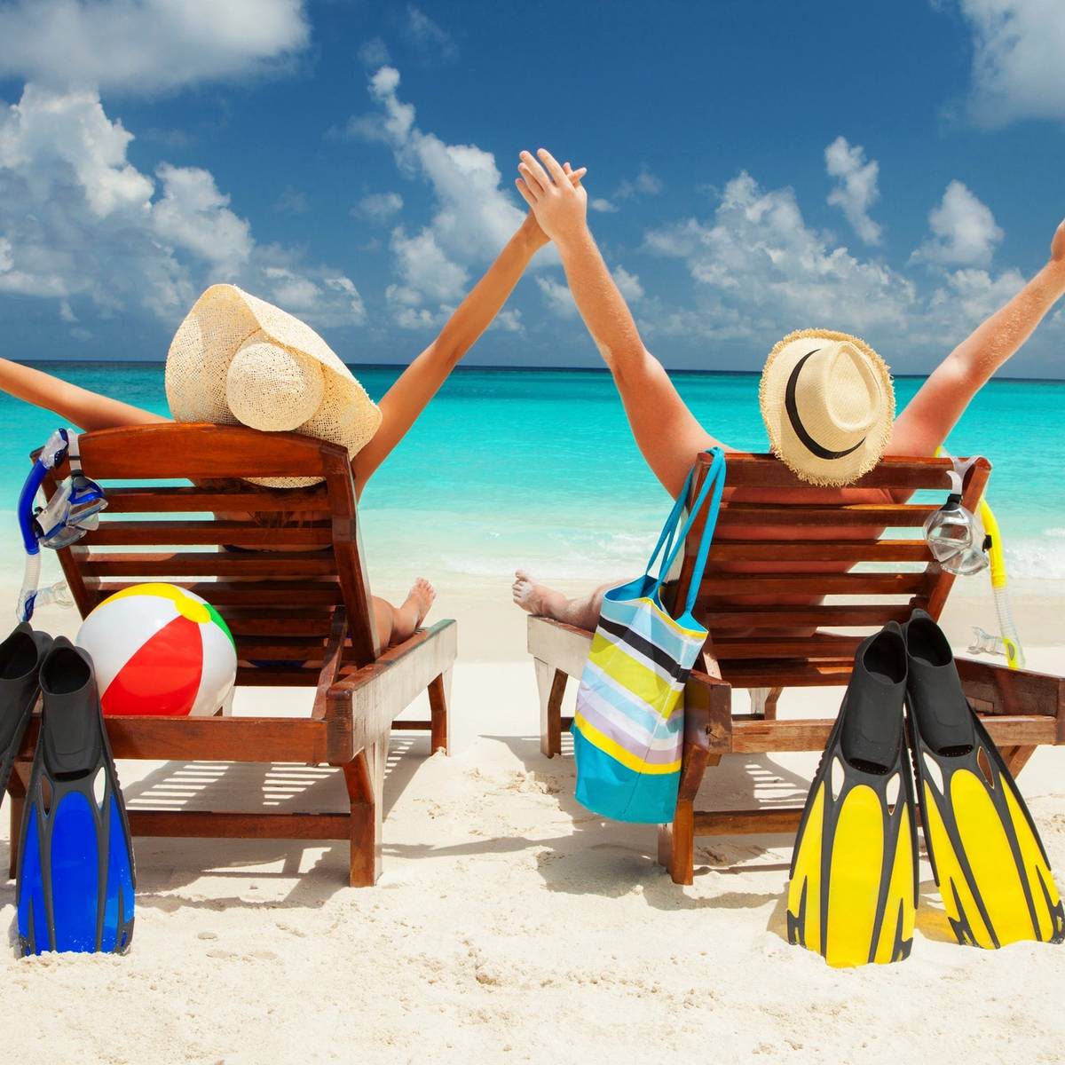 Two people relax on beach chairs by the ocean, wearing sun hats and flippers, with a beach ball nearby.