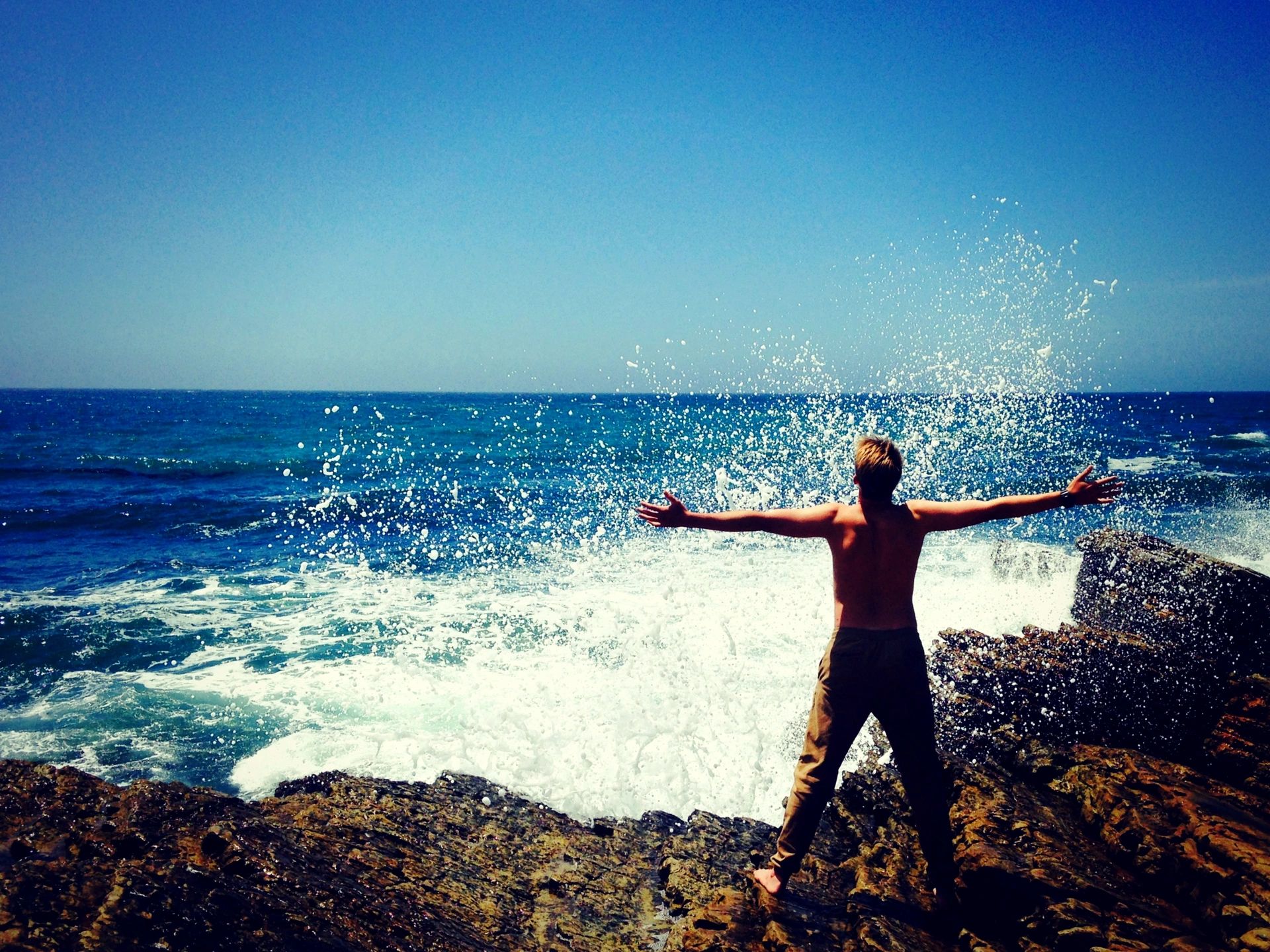 A person stands on rocks by the ocean, arms outstretched, as waves crash around them.